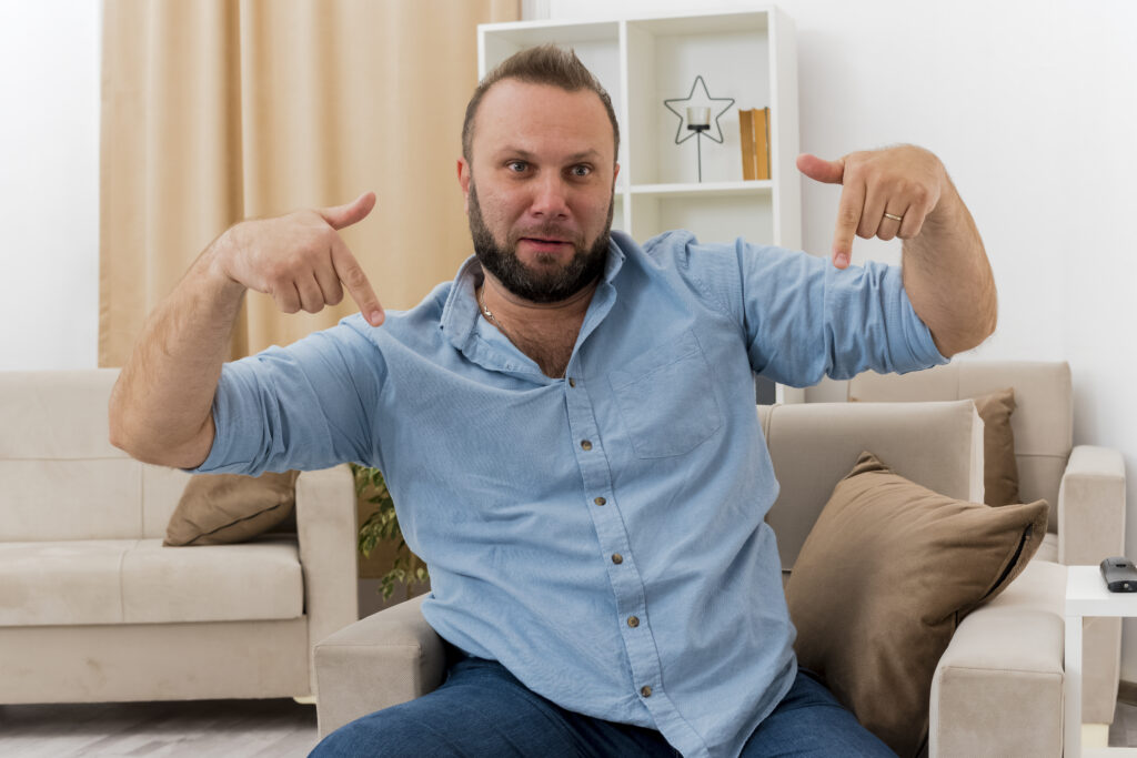 Man sitting on the couch with a worried expression and both hands up with index fingers pointing down.
